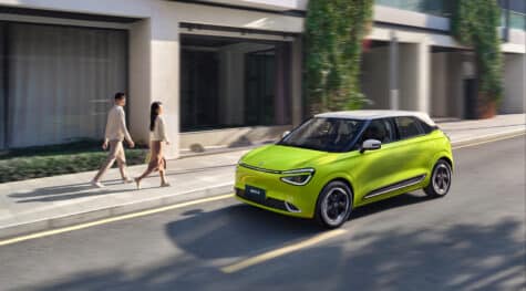 Geltonas miesto elektromobilis Dongfeng Box su baltu stogu važiuoja per miestą, vaizdas iš priekio ir šono